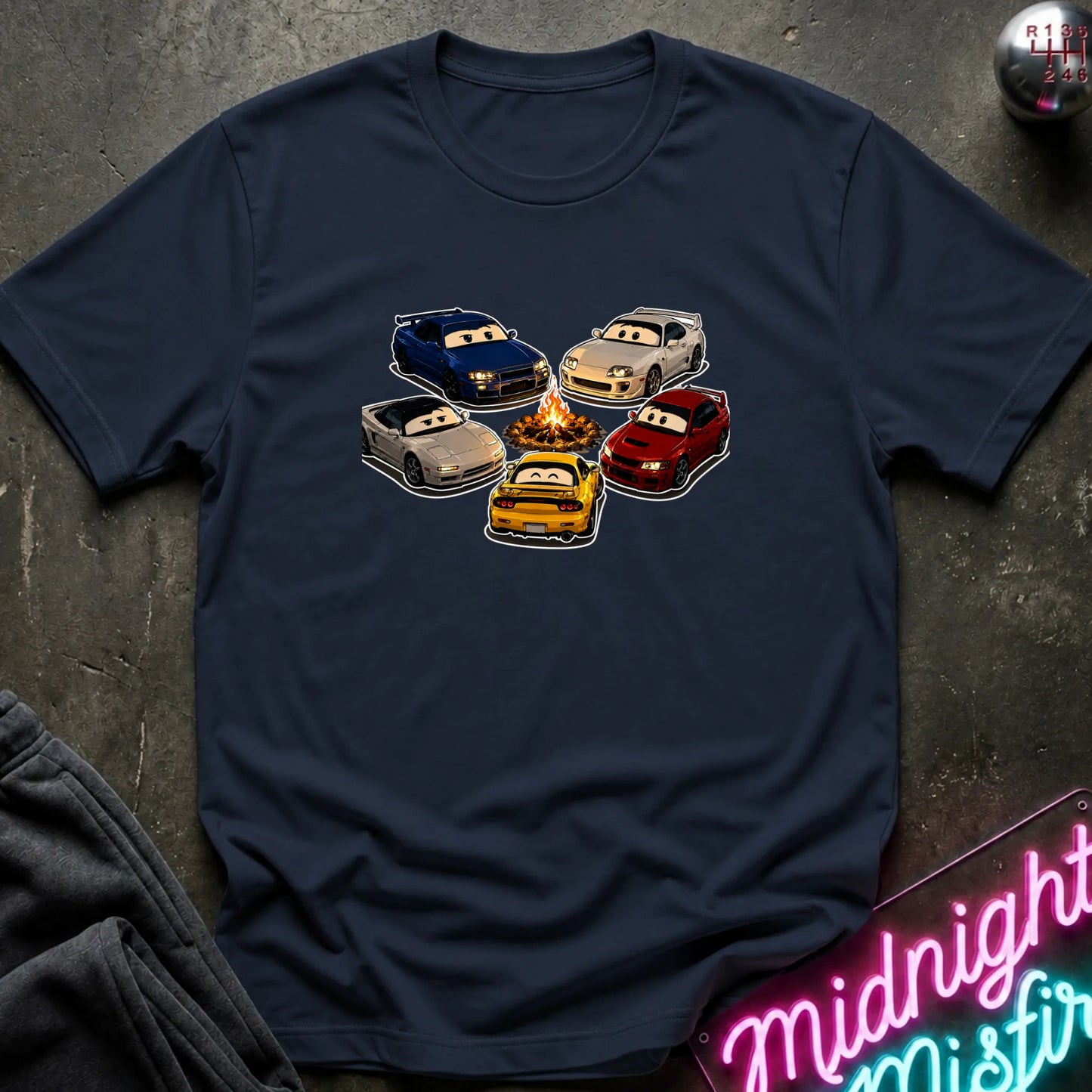 A navy blue short-sleeve t-shirt laid flat on a dark textured surface, featuring a graphic of cartoon race cars arranged around a campfire. The shirt is styled with a gray sweatshirt in the corner and a pink neon sign in the bottom right.