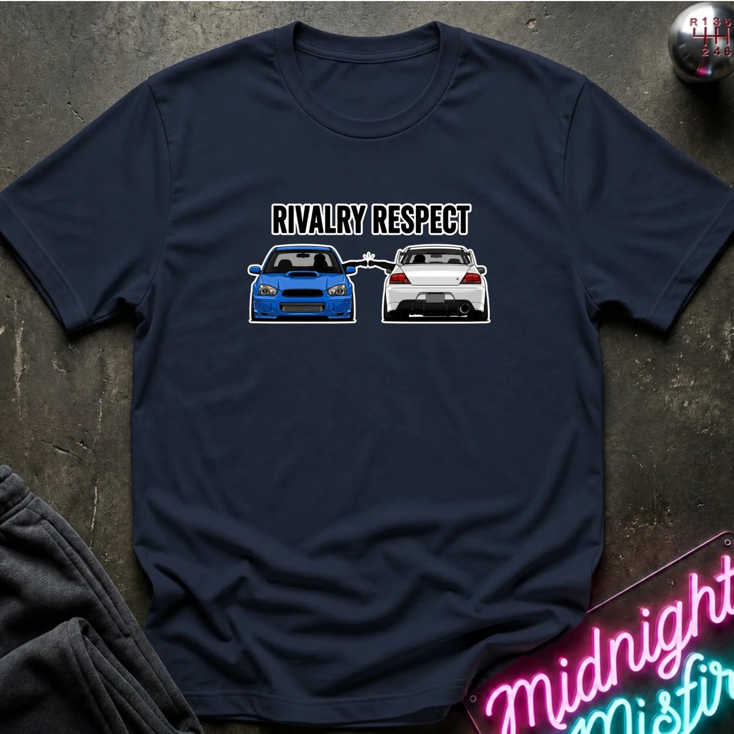 A navy blue short-sleeve graphic t-shirt laid flat on a dark textured surface. The graphic features the text 'RIVALRY RESPECT' above a blue and white car, 