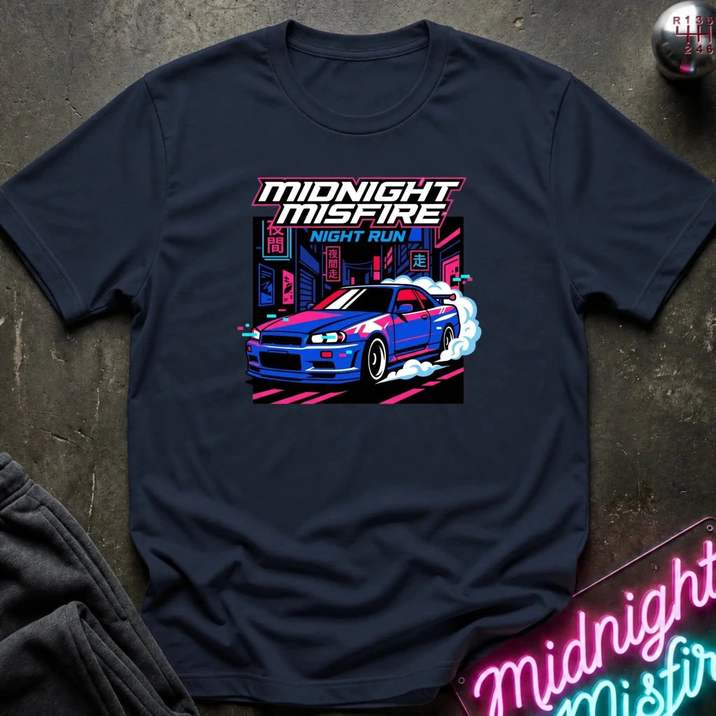 A navy blue crewneck t-shirt featuring a vibrant graphic of a blue and red sports car doing a burnout with neon city lights, under the text 'MIDNIGHT MISFIRE NIGHT RUN 2'. The shirt is laid flat on a dark textured surface with a matching neon sign in the corner.