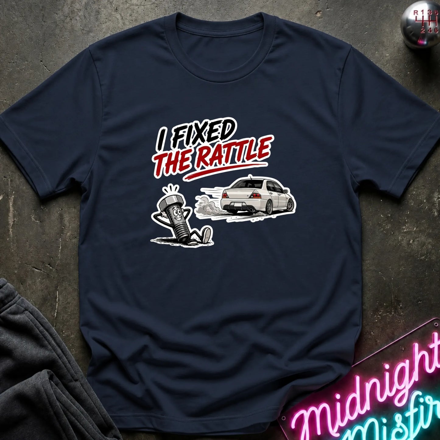 A navy blue graphic t-shirt laid flat on a dark textured surface. The graphic features the text 'I FIXED THE RATTLE' in white and red, above an illustration of a white sports car and a cartoon figure.