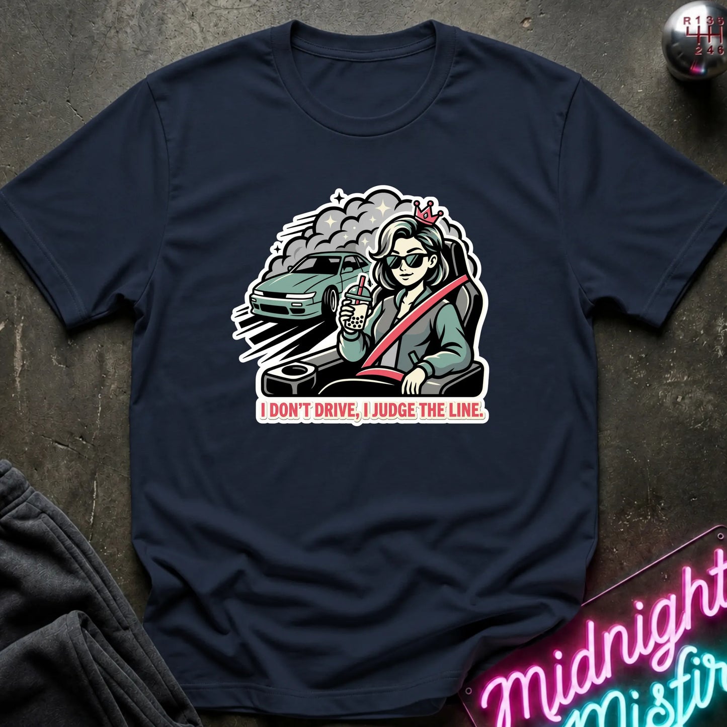 A navy blue short-sleeve t-shirt laid flat on a dark textured surface, featuring a colorful graphic of a woman in a car with the text 'I DON'T DRIVE, I JUDGE THE LINE.' A neon sign and folded dark fabric are partially visible around the shirt.