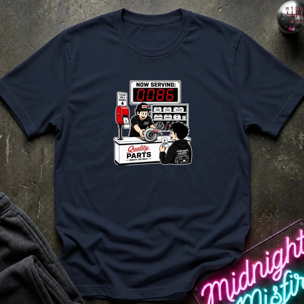 A navy blue crewneck t-shirt laid flat on a dark textured surface, featuring a retro-style graphic of a mechanic working at a 'Quality Parts' counter with a red digital display showing '0086'.
