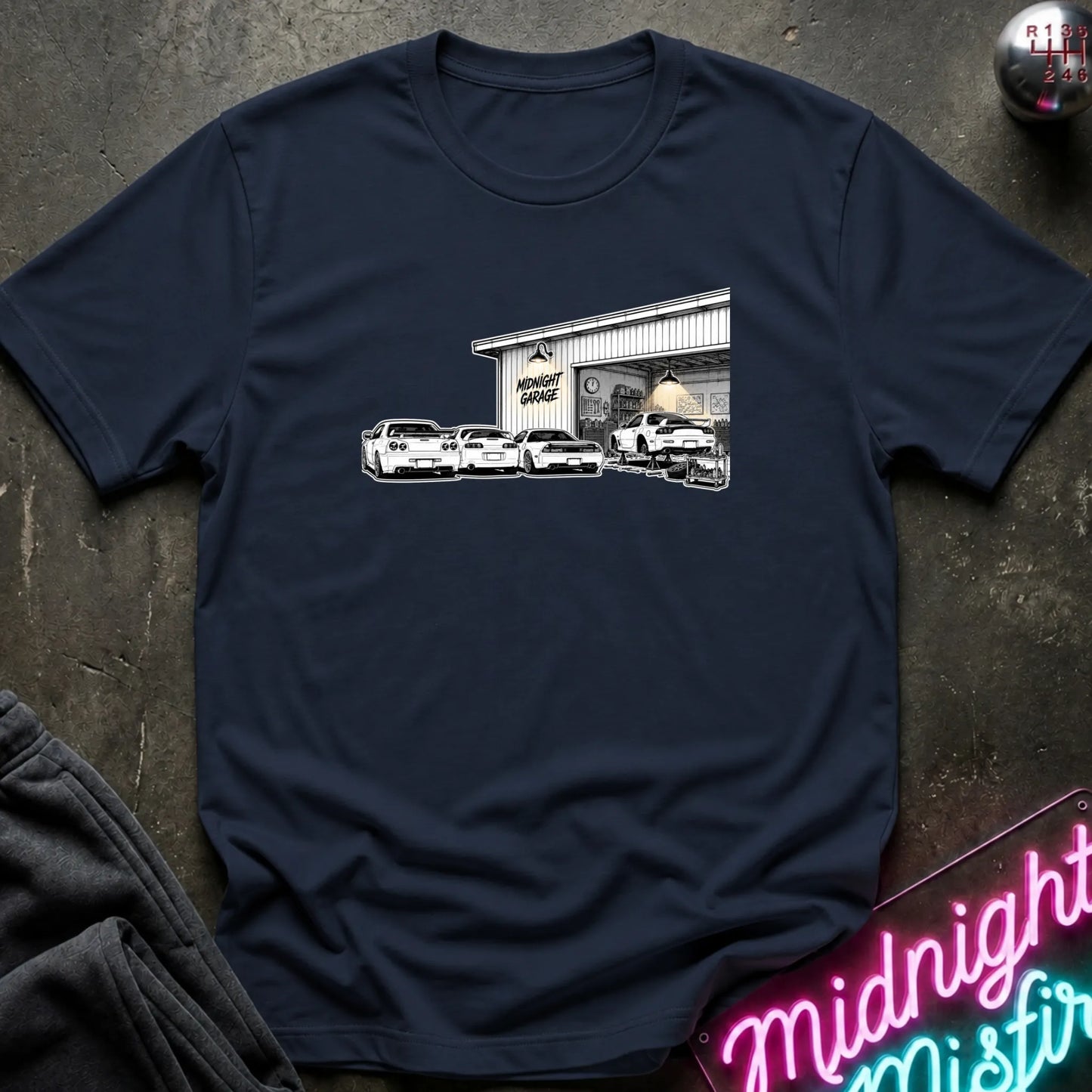 A navy blue short-sleeve t-shirt laid flat on a dark textured surface, featuring a black and white graphic print of a garage with several cars in front. 