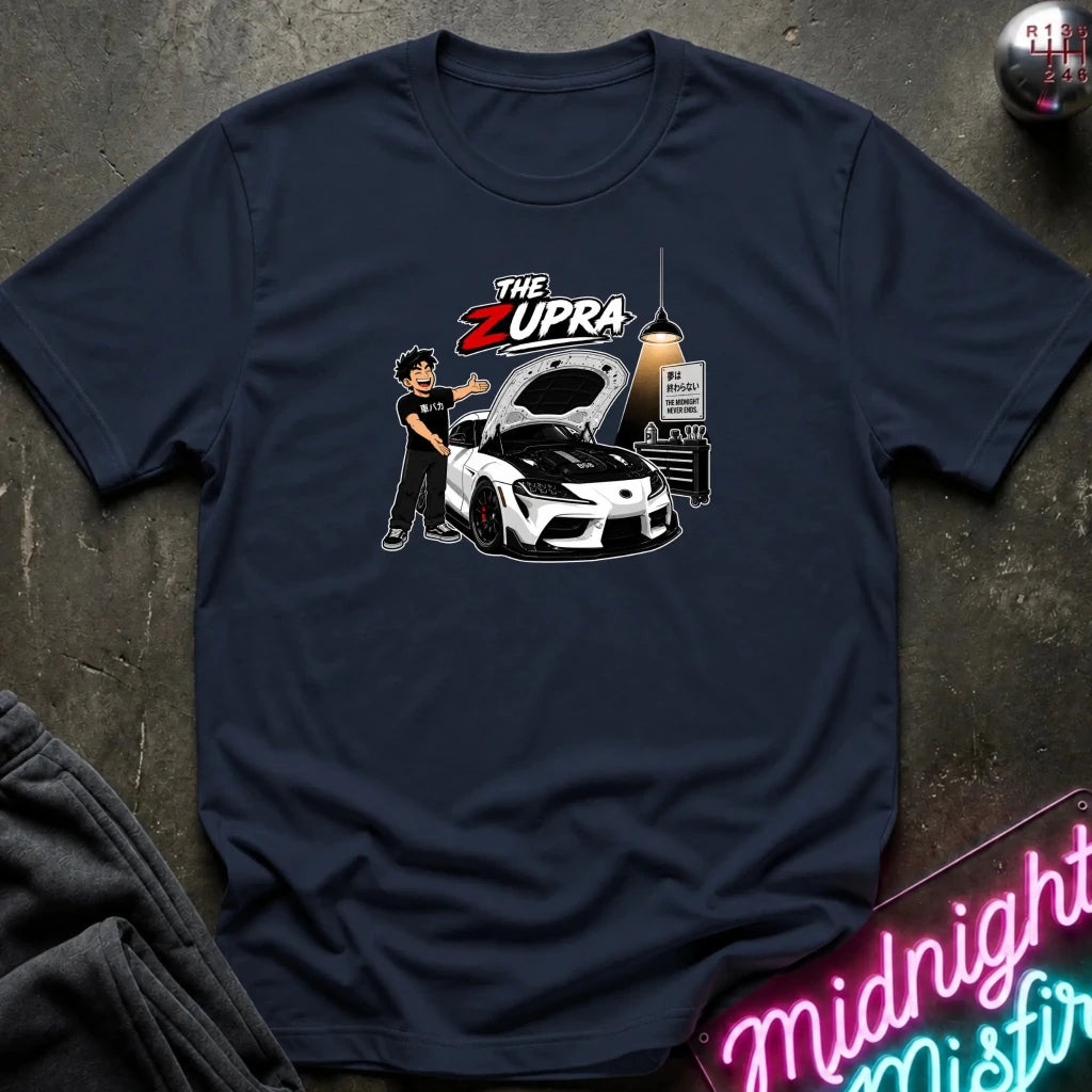 A navy blue short-sleeve graphic t-shirt laid flat on a dark textured surface. The graphic features a cartoon illustration of a man standing next to a white sports car with its hood open, under the text 'THE ZUPRA'.