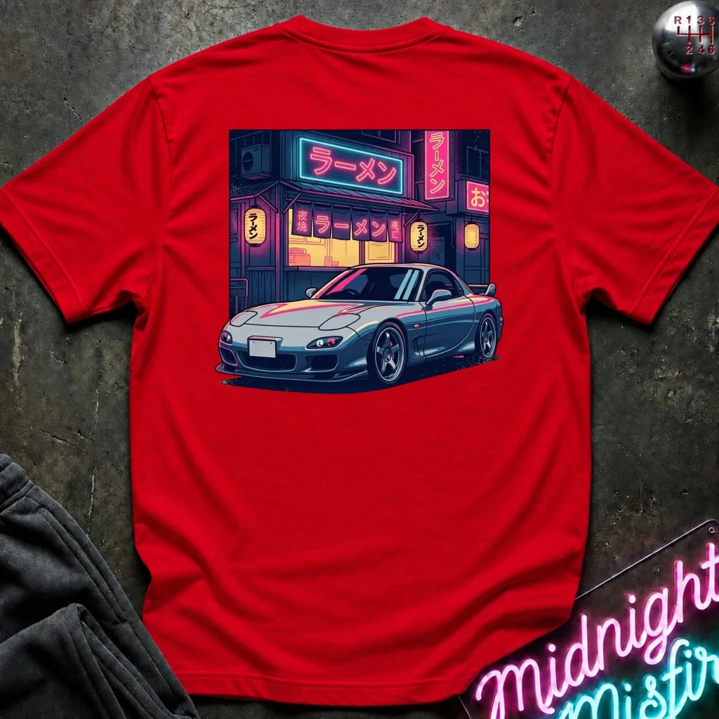 A bright red short-sleeve t-shirt laid flat on a dark textured surface, featuring a large graphic print on the back of a silver sports car in front of a neon-lit ramen shop. 
