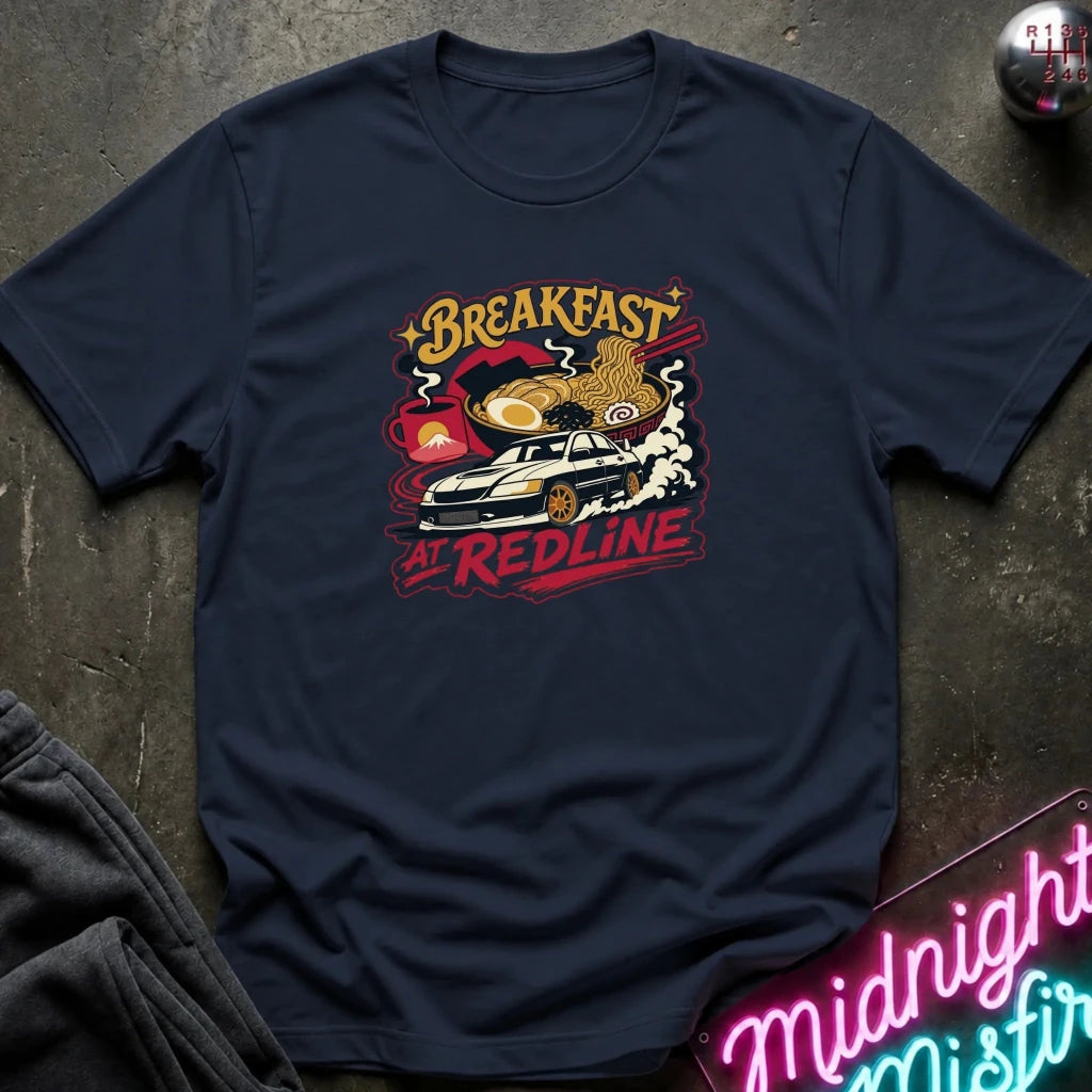 A navy blue short-sleeve graphic T-shirt laid flat on a dark textured surface. The shirt features a colorful retro-style chest print with the words 'Breakfast at Redline'