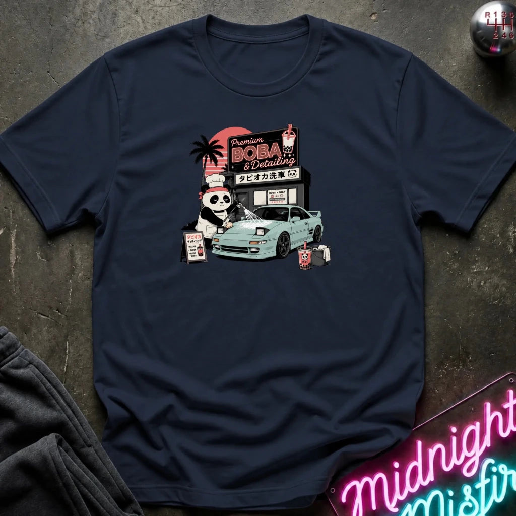 navy short-sleeve graphic t-shirt laid flat on a dark textured surface. The graphic features a cartoon panda washinga light blue car