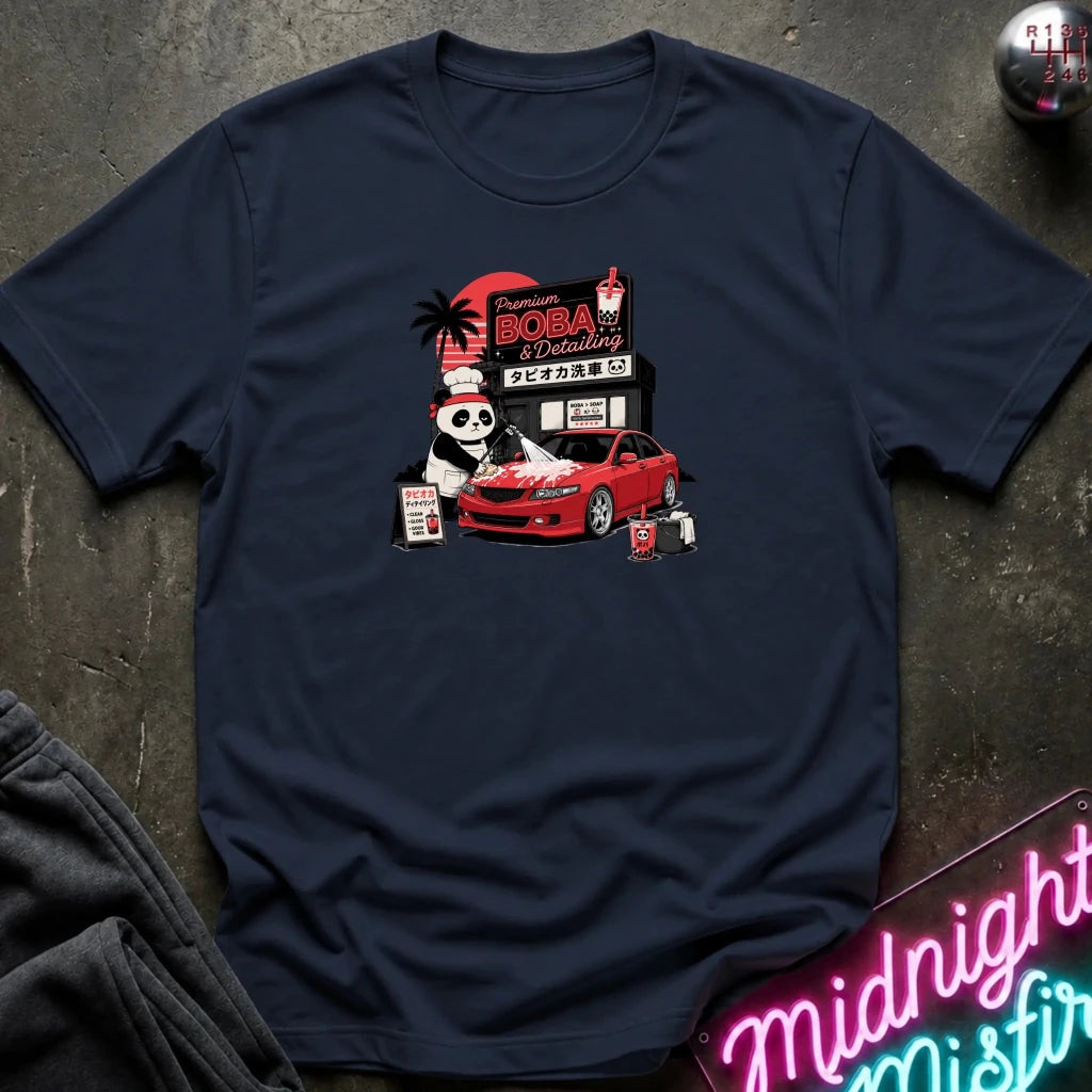 navy short-sleeve graphic t-shirt laid flat on a dark textured surface. The graphic features a cartoon panda wearing a hat next to a red sports car, with a sign reading 'Midnight Boba'.