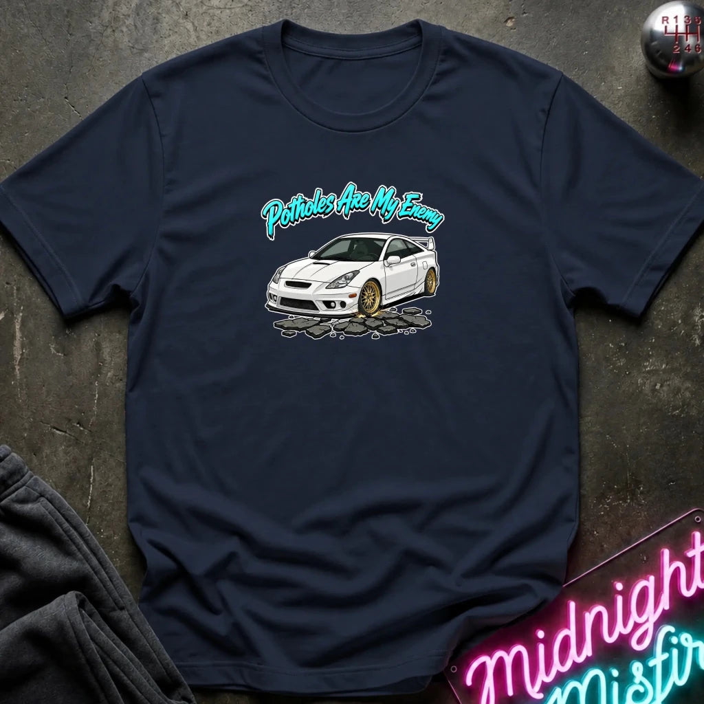 A navy blue graphic t-shirt laid flat on a dark textured surface. The shirt features a white sports car with gold wheels and the text 'Pistons Are My Enemy' in a teal cursive font.