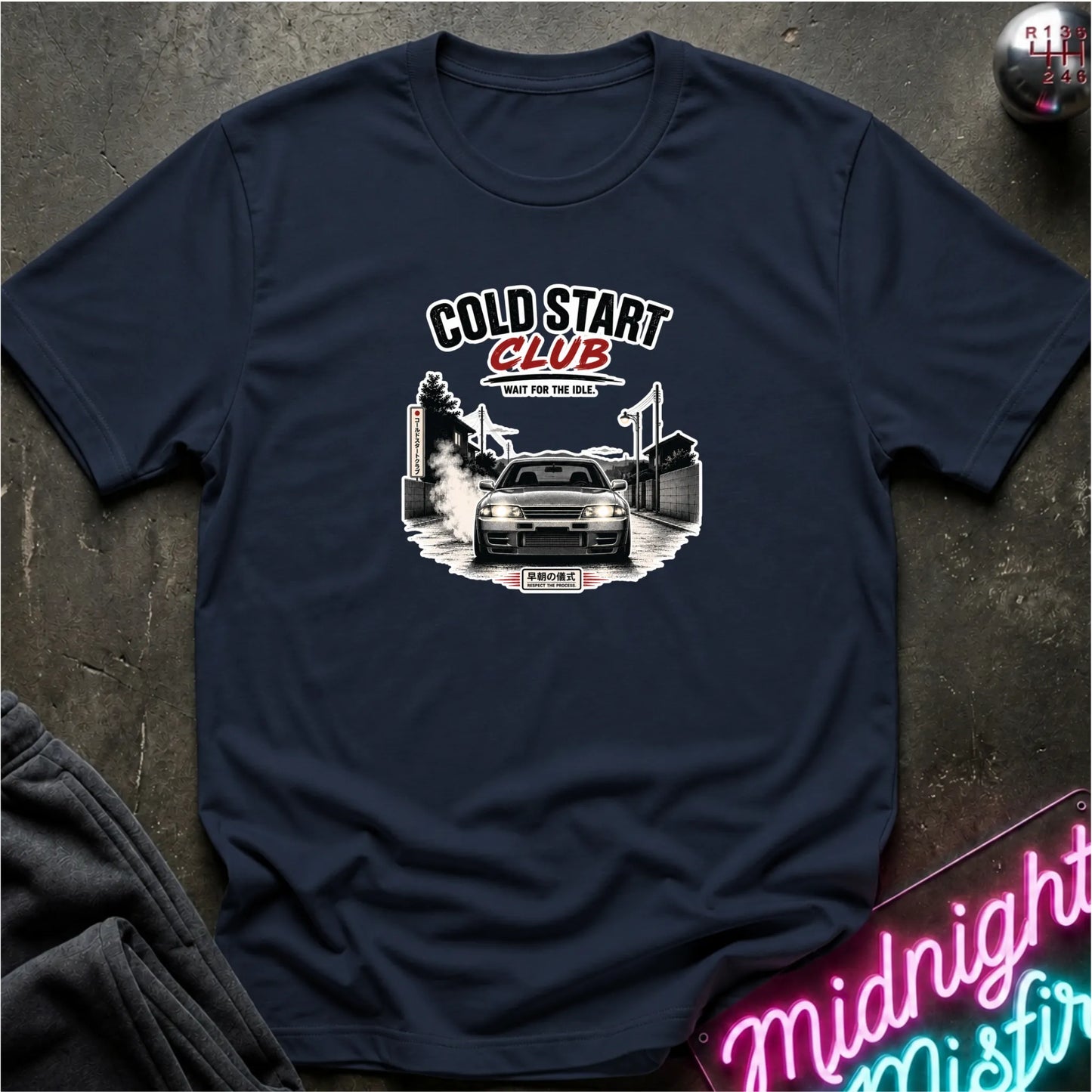 A navy blue short-sleeve t-shirt with a 'Cold Start Club' car graphic, laid flat on a dark textured surface with a neon sign and dark clothing in the background.