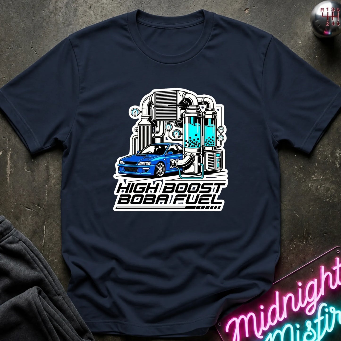 A navy blue short-sleeve t-shirt laid flat on a dark textured surface, featuring a graphic of a blue sports car in front of a high-tech turbocharger illustration with the text 'HIGH BOOST BOBAI FUEL'.
