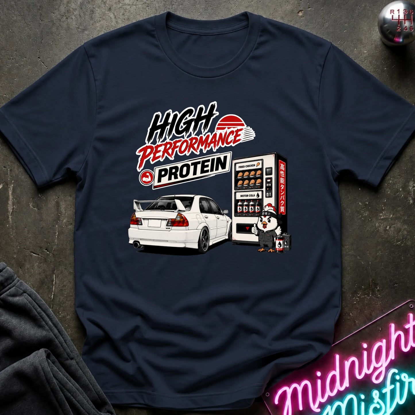 A navy blue graphic t-shirt laid flat on a dark textured surface. The shirt features a humorous graphic combining a car and a vending machine with the text 'HIGH PERFORMANCE PROTEIN'.