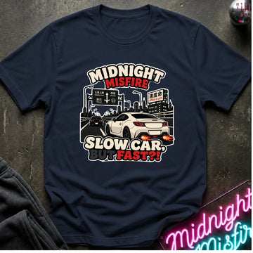 A navy blue graphic t-shirt laid flat on a dark textured surface. The shirt features a graphic of a white sports car driving slowly in front of city signs, with the text 'MIDNIGHT MISFIRE' and 'SLOW CAR.