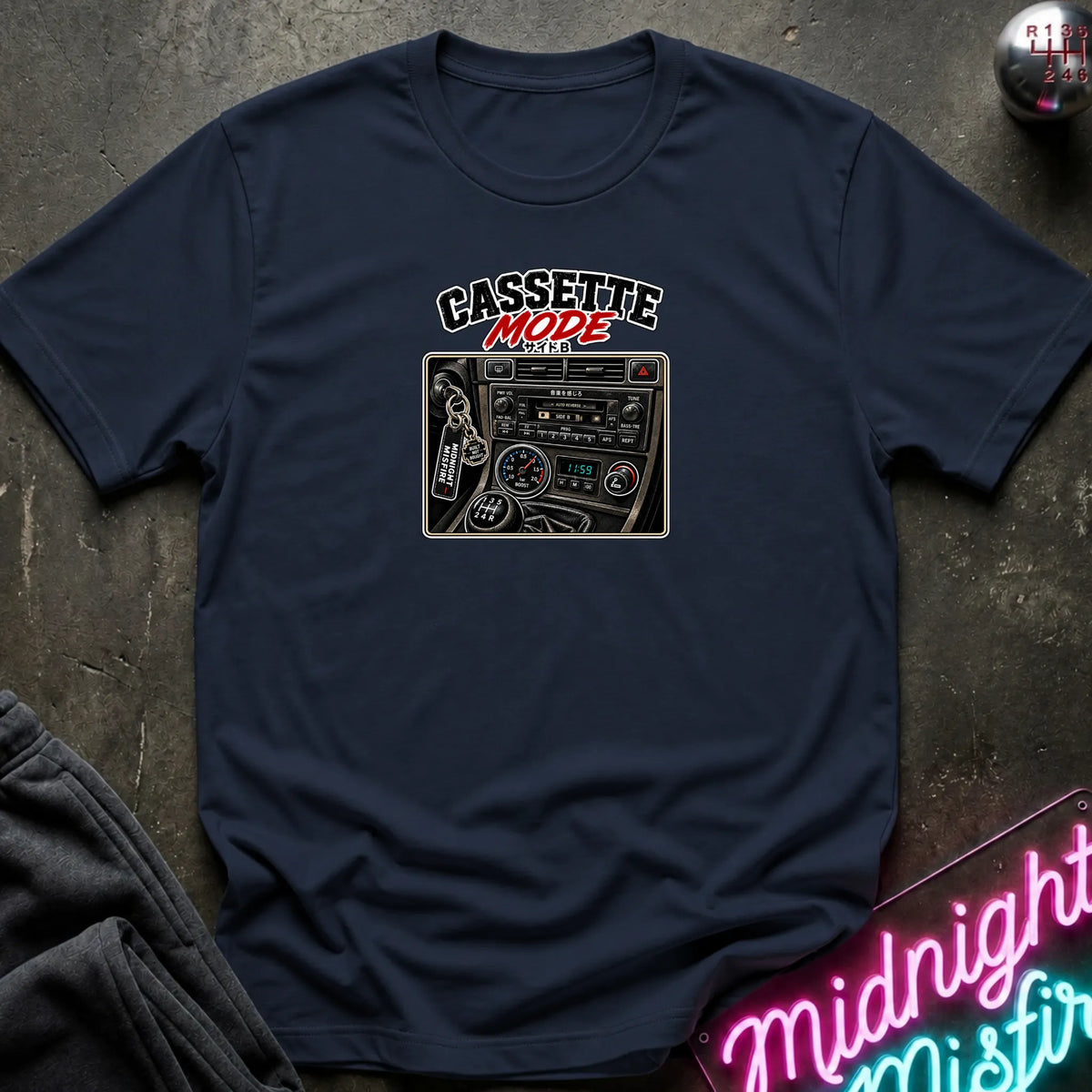 A navy short-sleeve t-shirt laid flat on a dark textured surface, featuring a graphic print of a cassette radio with the text 'CASSETTE MODE' in black and red. A neon sign and dark pants are partially visible as props.