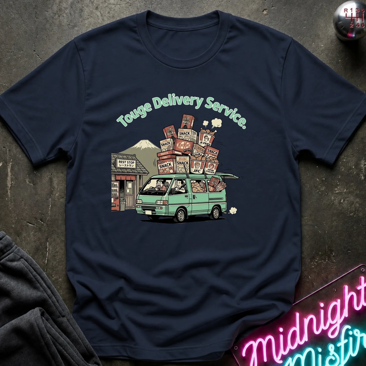 A navy blue  short-sleeve t-shirt with a graphic print of a green van carrying stacked cardboard boxes under the text 'Touge Delivery Service.' The shirt is laid flat on a dark textured surface with a neon sign and dark fabric partially visible at the edges.