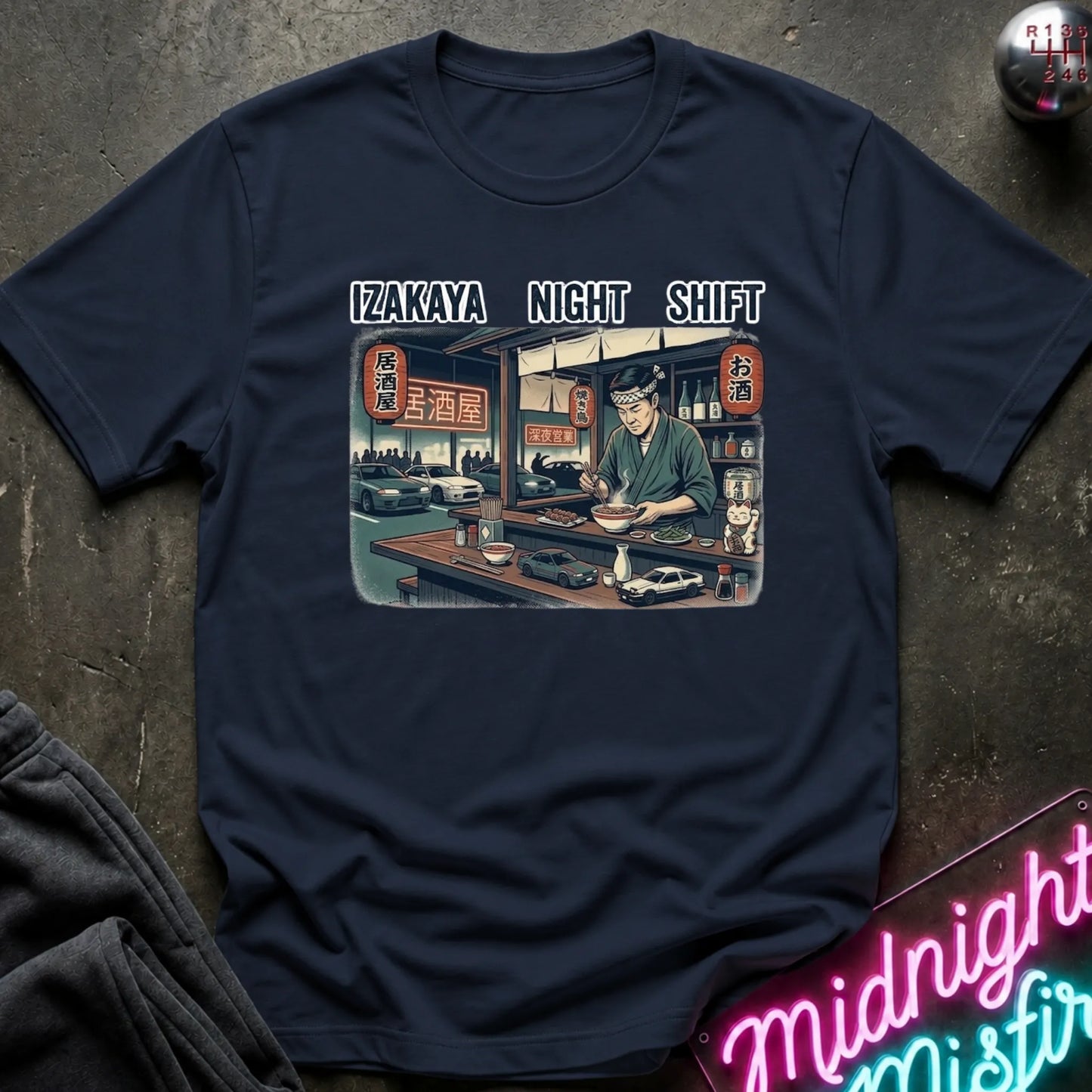 A navy blue graphic t-shirt laid flat on a dark textured surface. The shirt features a central rectangular illustration of a Japanese izakaya bar scene with a bartender, and the text 'IZAKAYA NIGHT SHIFT' printed above it.