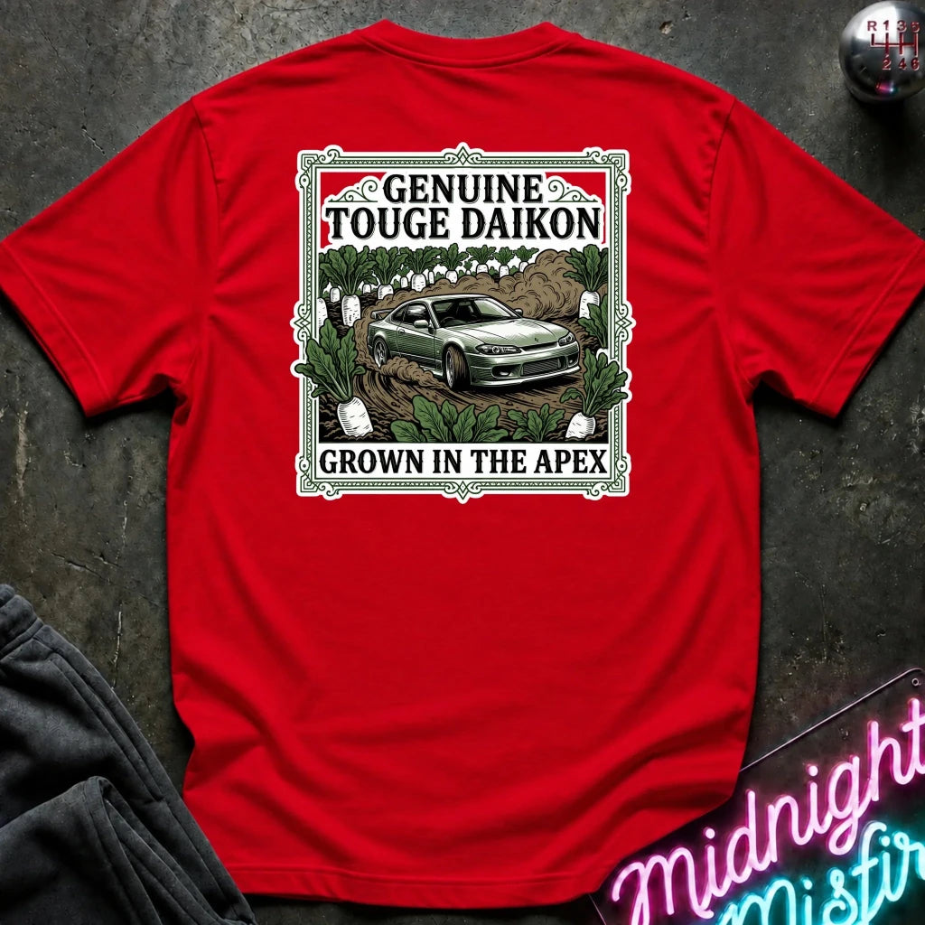 A red short-sleeve graphic T-shirt laid flat on a dark textured surface. The shirt features a rectangular vintage-style print with the words 'Genuine Touge Daikon' and 'Grown in the Apex' surrounding an illustration of a green car in a field.