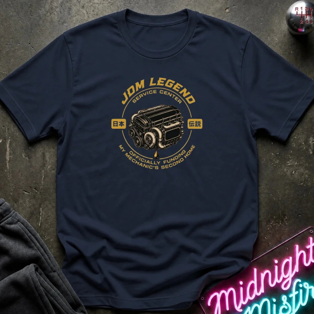 navy graphic t-shirt laid flat on a dark textured surface. The shirt features a vintage-style yellow and black engine graphic with the text 'JDM LEGEND' and 'MY SPECIALTY FUNDS MECHANIC'S SECOND HAND'.