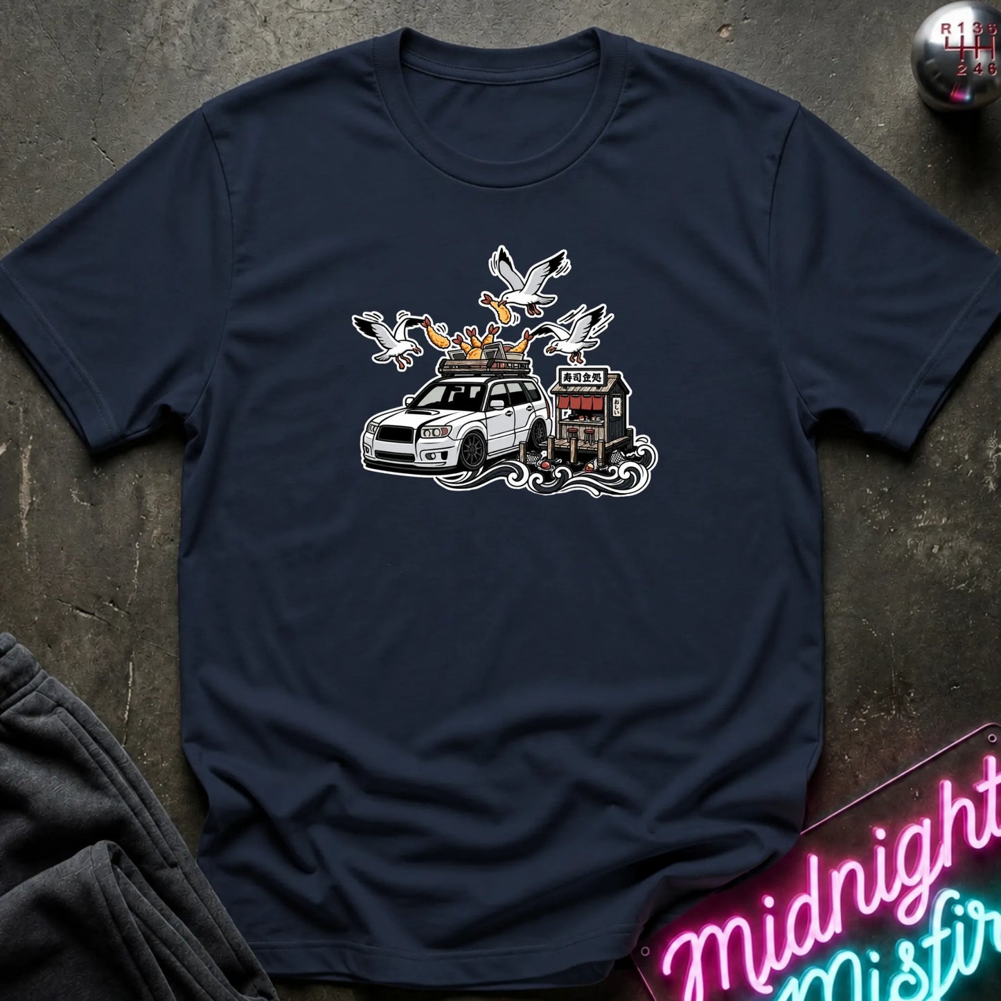 A navy blue crewneck t-shirt laid flat on a dark textured surface, featuring a graphic print of a white station wagon with a roof rack carrying seagulls and a small building. A neon sign reading 'Midnight Misfire' is visible in the bottom right corner.