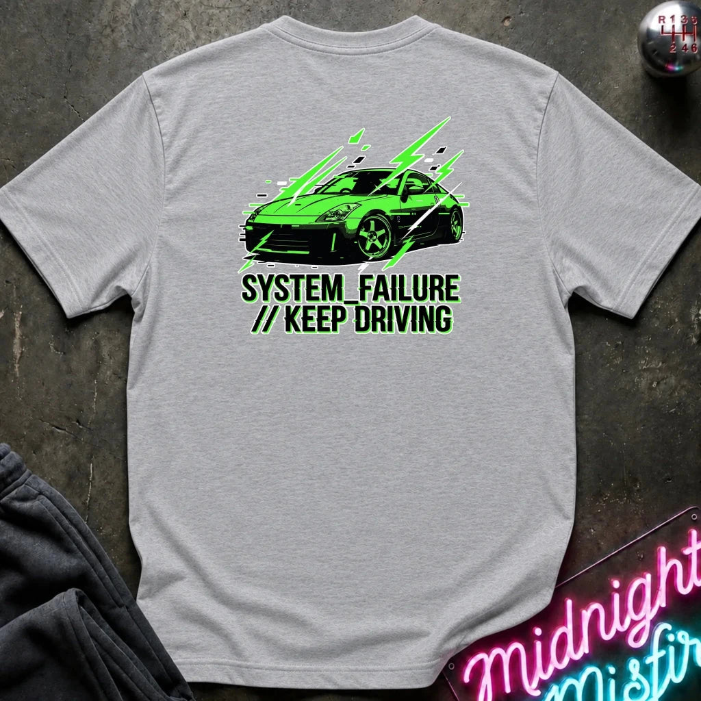 gray short-sleeve t-shirt laid flat on a dark textured surface, featuring a graphic of a green sports car with text 'SYSTEM FAILURE // KEEP DRIVING' in green. A neon sign and dark clothing are partially visible as props.