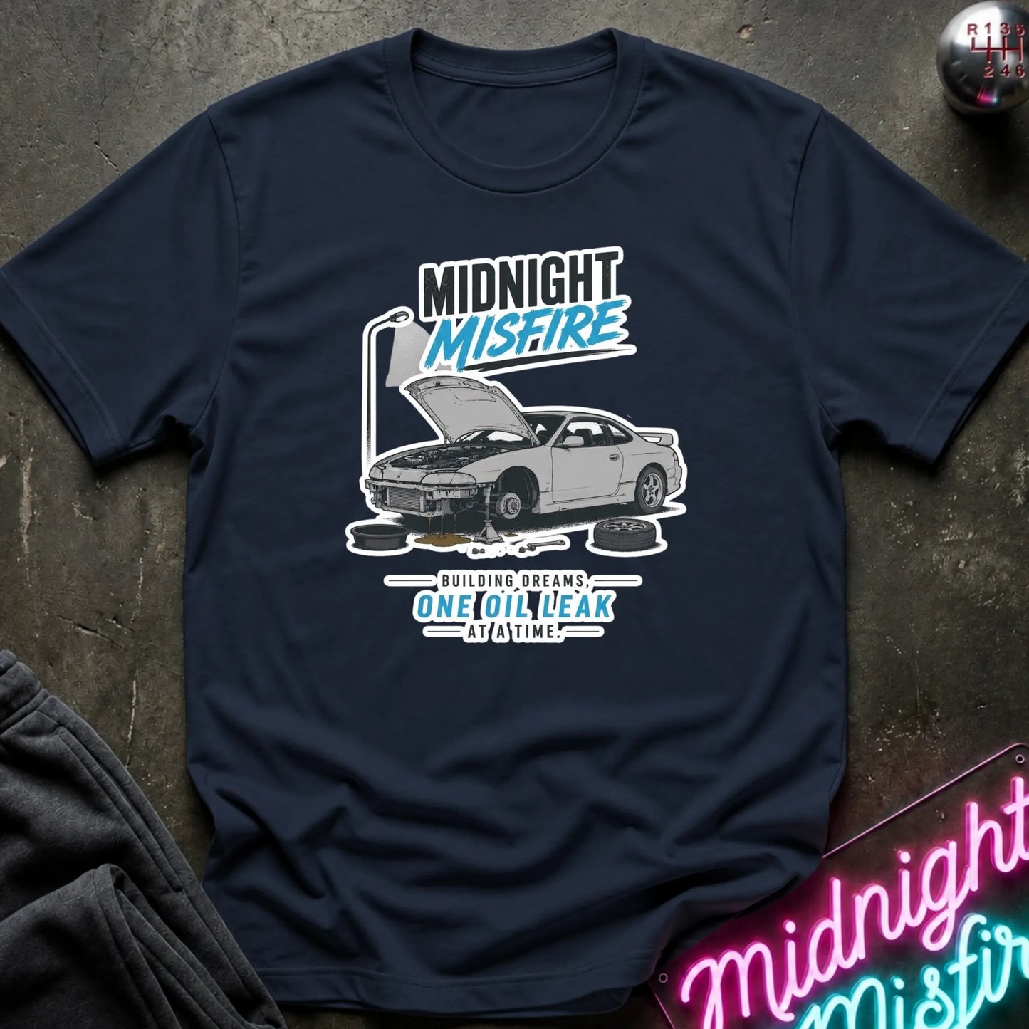 A navy blue short-sleeve graphic T-shirt laid flat on a dark textured surface. The shirt features a printed design of a silver car with its hood open, accompanied by the text 'MIDNIGHT MISFIRE' and 'BUILDING DREAMS.