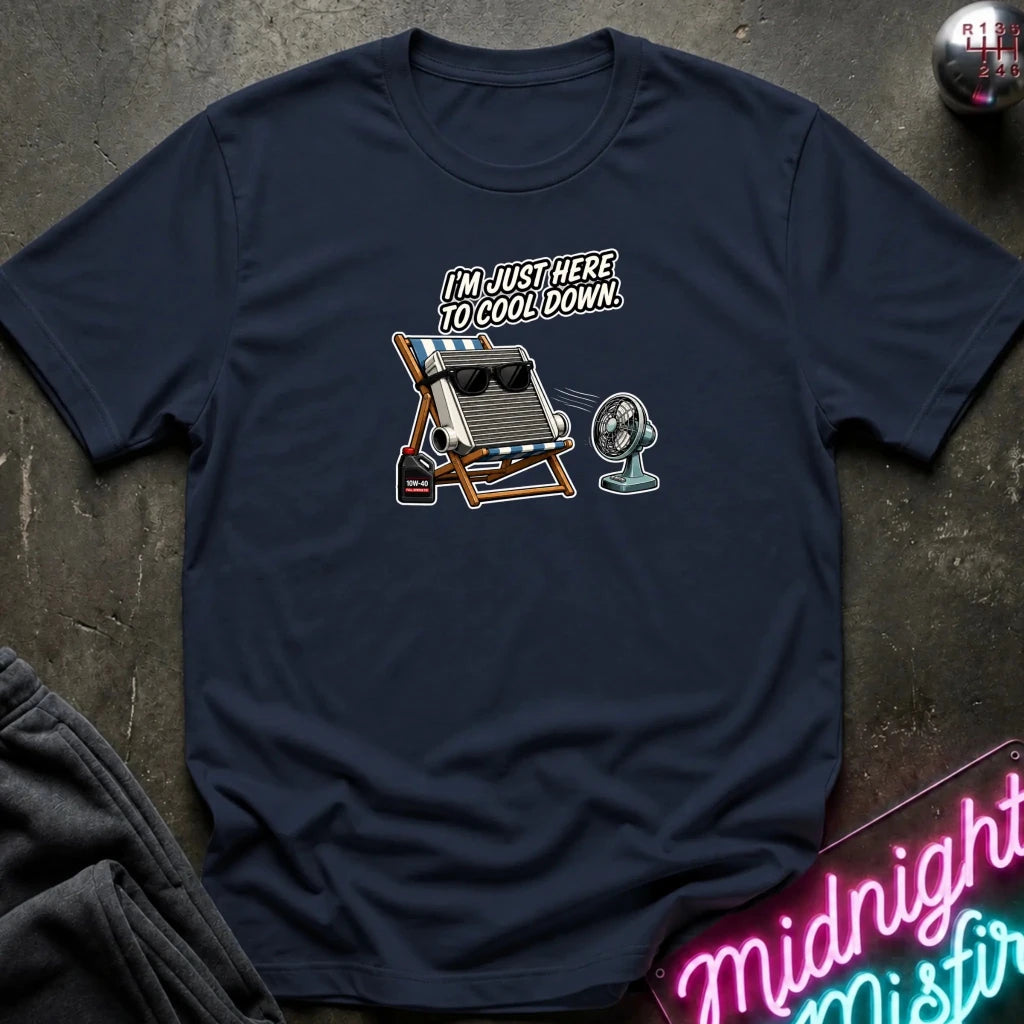 A navy blue short-sleeve t-shirt with a graphic of a vintage fan, a beach chair with sunglasses, and the text 'I'M JUST HERE TO COOL DOWN!' laid flat on a dark textured surface.