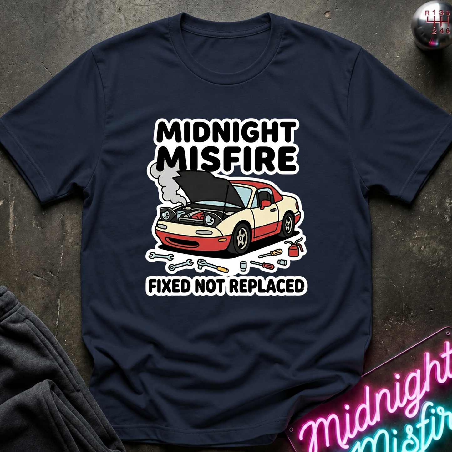 A navy blue graphic t-shirt laid flat on a dark textured surface. The shirt features a cartoon illustration of a beige and red sports car with its hood open, surrounded by wrenches and smoke, with the text 'MIDNIGHT MISFIRE' above and 'FIXED NOT REPLACED' below.