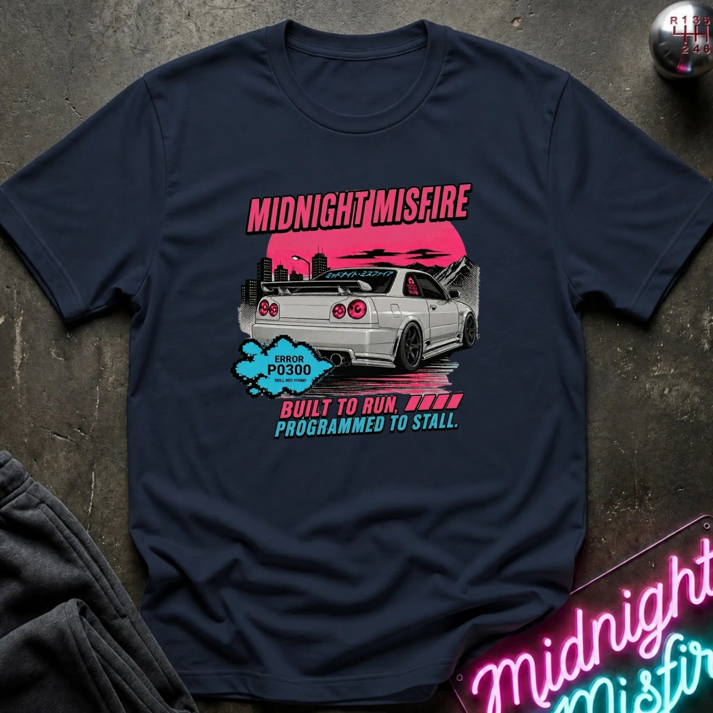 A navy short-sleeve graphic t-shirt laid flat on a dark textured surface. The graphic features a white sports car with a pink sunset, the text 'MIDNIGHT MISFIRE' in pink.