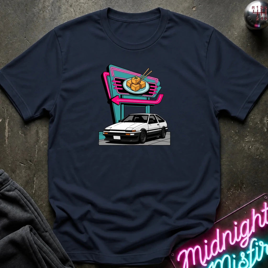 A navy blue short-sleeve t-shirt laid flat on a dark textured surface, featuring a colorful graphic print of a white hatchback parked in front of a neon-style food sign. 