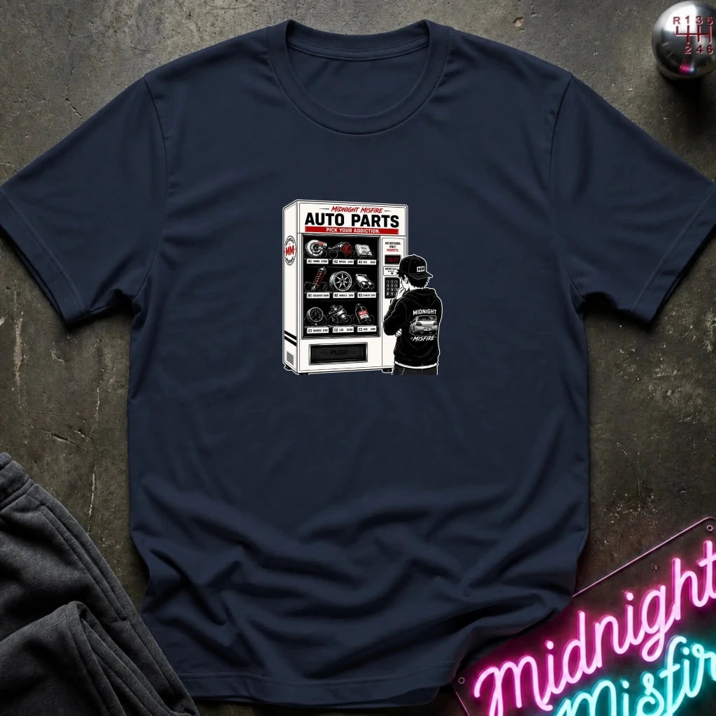 A navy blue crewneck t-shirt laid flat on a dark textured surface, featuring a graphic print of a person in a black hoodie standing in front of an 'Auto Parts' vending machine. 