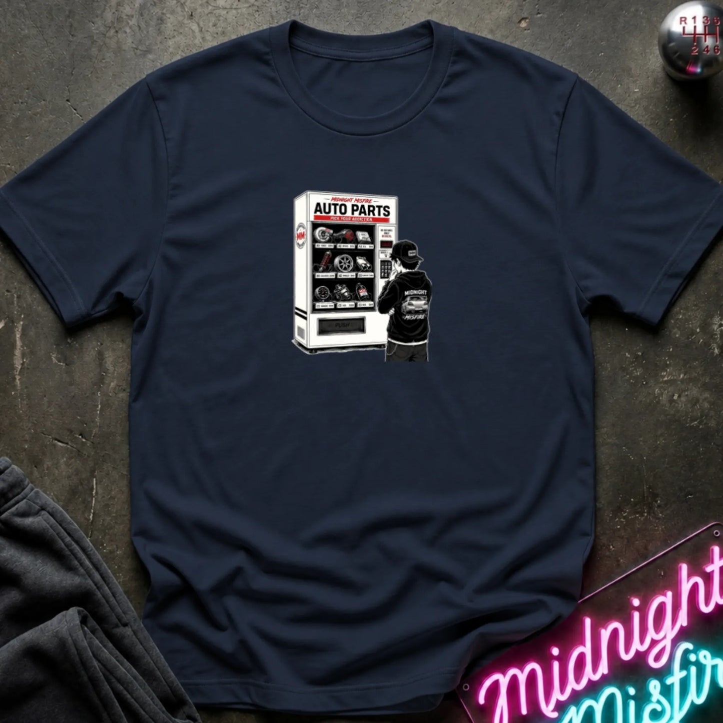A navy short-sleeve t-shirt laid flat on a dark textured surface, featuring a graphic print of a vending machine and owner deciding.