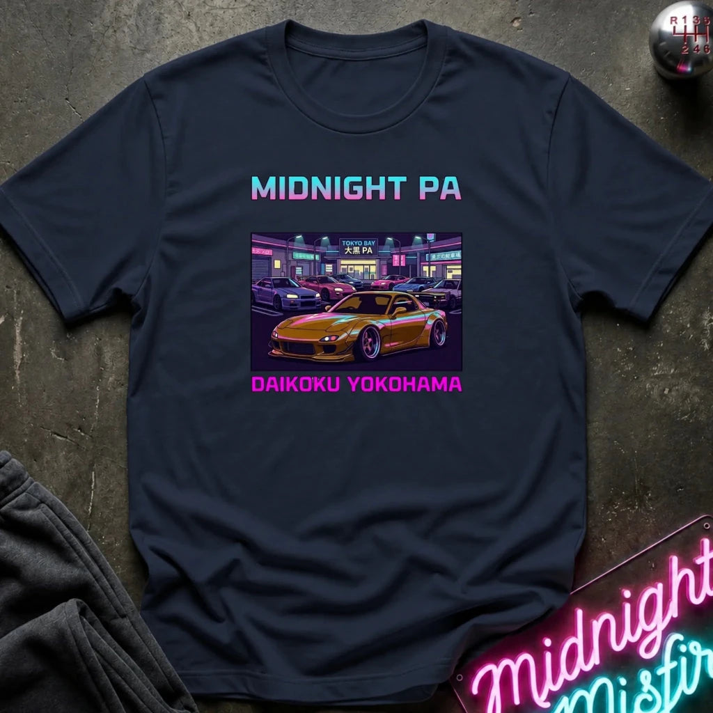 A navy blue graphic t-shirt laid flat on a dark textured surface. The shirt features a colorful retro-style print of a yellow sports car in a neon-lit parking lot at night, with the text 'MIDNIGHT PA' above and 'DAIKOKU YOKOHAMA' below in pink.