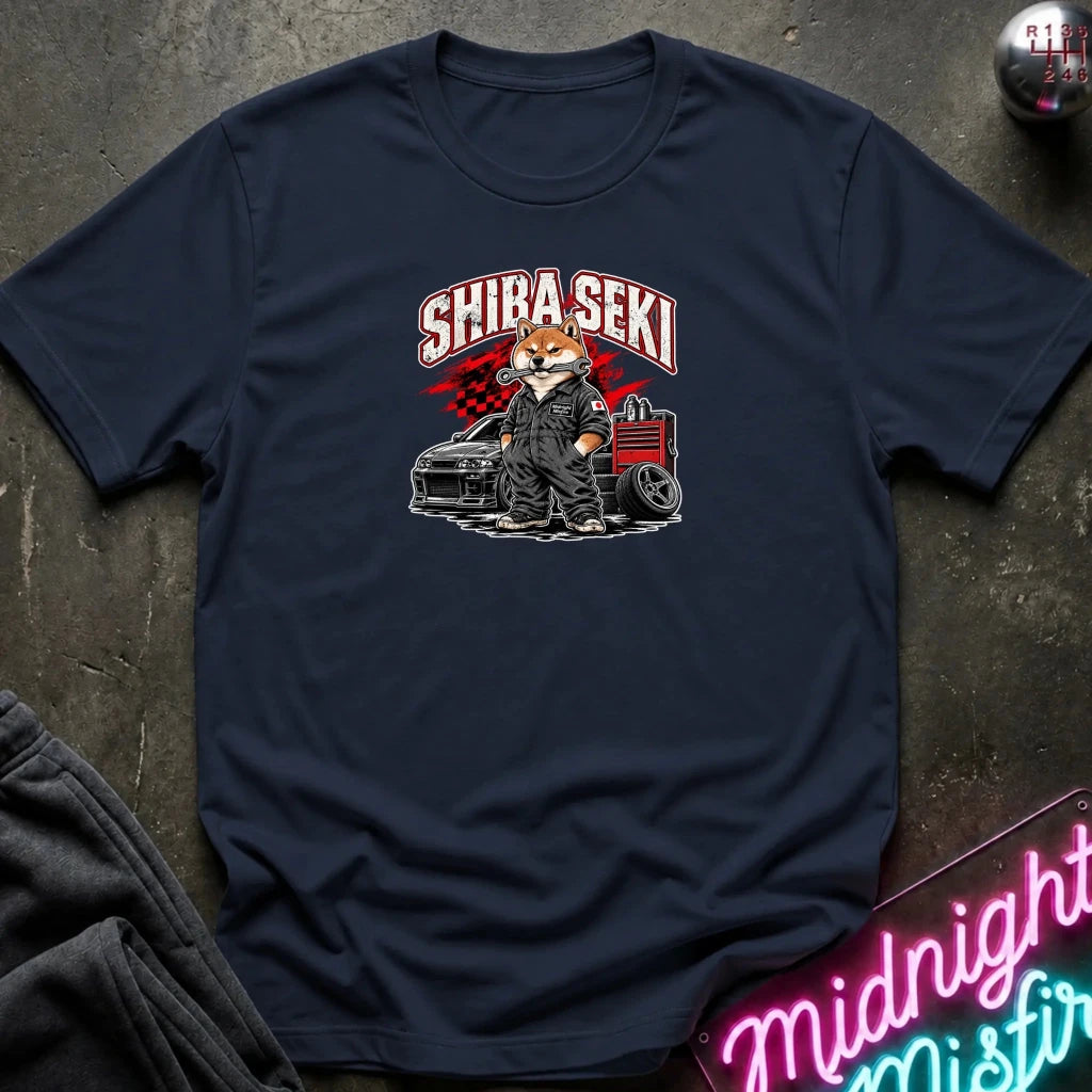 A navy blue graphic t-shirt laid flat on a dark textured surface. The shirt features a graphic of a fox character in a suit standing next to a car and tool chest, with the text 'SHIBA SEKI' above.
