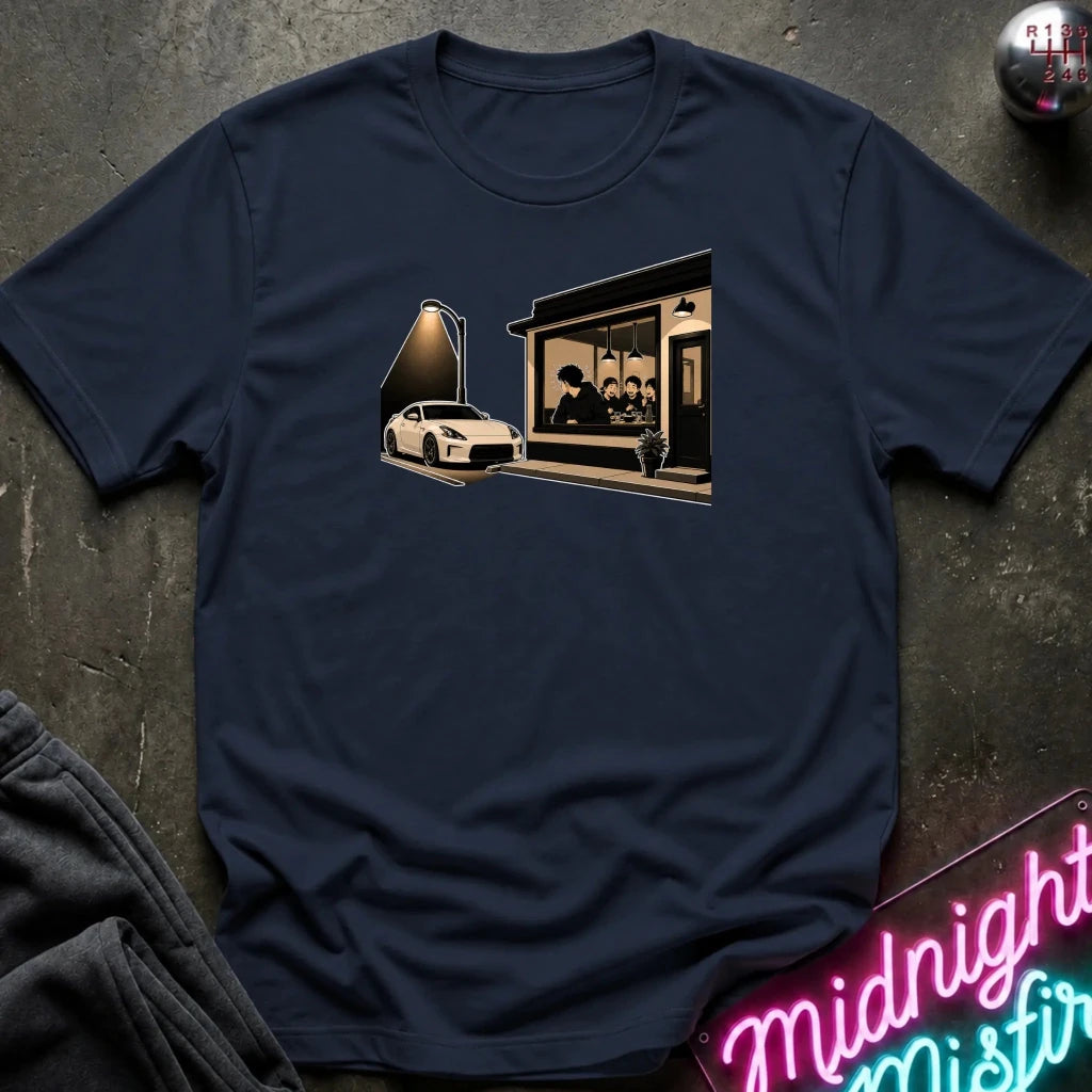 A navy blue short-sleeve t-shirt laid flat on a dark textured surface, featuring a graphic print of a white sports car parked outside a dimly lit cafe with a neon sign in the foreground. 