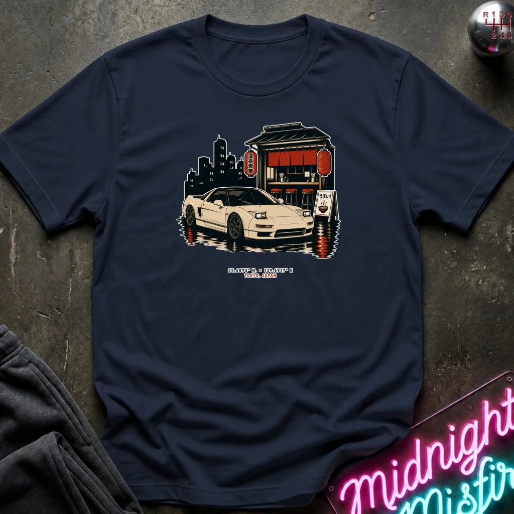A navy blue graphic t-shirt laid flat on a dark textured surface. The graphic features a white sports car parked in front of a Japanese-style building with red lanterns and a city skyline silhouette.