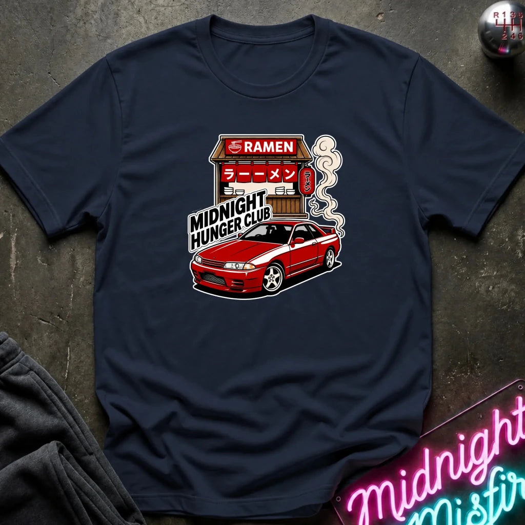 A navy blue graphic t-shirt laid flat on a dark textured surface. The graphic features a red sports car in front of a ramen shop with the text 'RAMEN', 'MIDNIGHT HUNGER CLUB', and 'Midnight Hunger Club'.