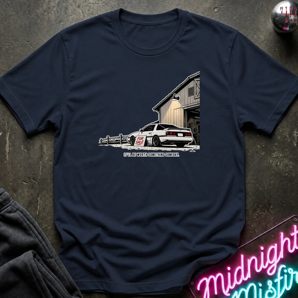 A navy blue short-sleeve t-shirt laid flat on a dark textured surface, featuring a graphic print of a white sports car parked in front of a barn with the text.