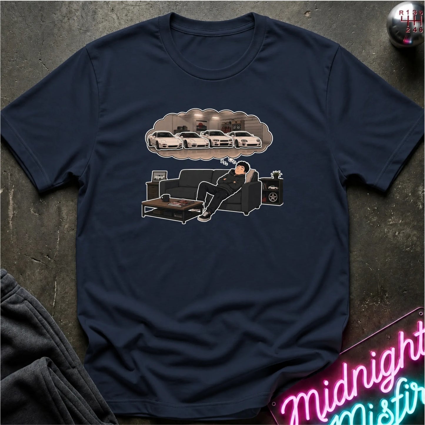 A navy blue graphic t-shirt laid flat on a dark textured surface. The graphic shows a man lounging on a couch with a thought bubble above him containing white cars.