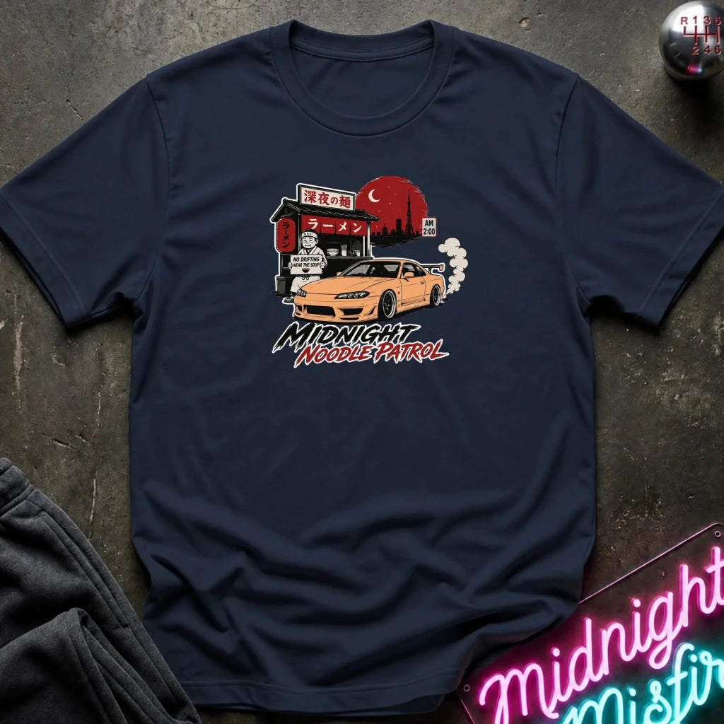 navy crewneck t-shirt featuring a graphic print of a yellow Nissan Silvia S15 drift car in front of a red moon and Japanese storefront, with the text 'MIDNIGHT Noodle PATROL'. The shirt is laid flat on a dark textured surface with a neon sign and dark pants partially visible.