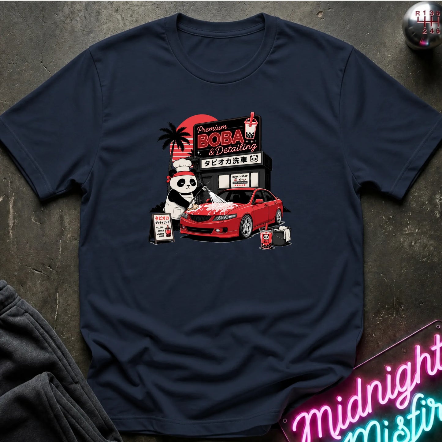 A navy blue short-sleeve t-shirt with a colorful graphic print of a panda working at a car detailing shop next to a red sports car, laid flat on a dark textured surface.