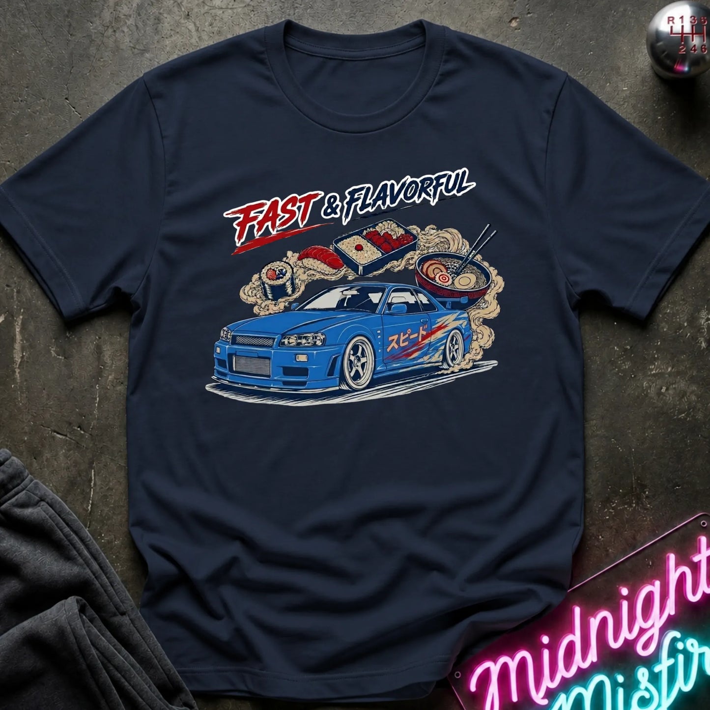 A navy blue graphic t-shirt laid flat on a dark textured surface. The shirt features a colorful illustration of a blue sports car with exhaust flames and ramen bowls, accompanied by the text 'FAST & FLAVORFUL' in red and white.