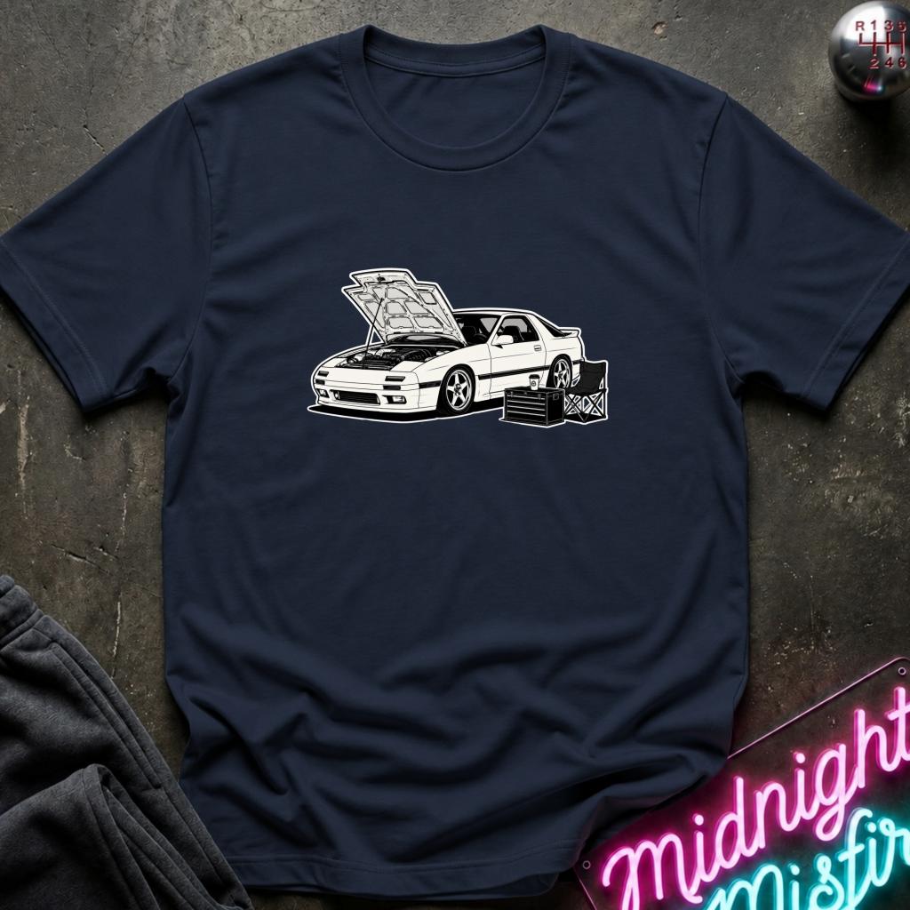 A navy blue short-sleeve t-shirt with a white graphic of a sports car with its hood open, laid flat on a dark textured surface with a neon sign and clothing props in the corners.