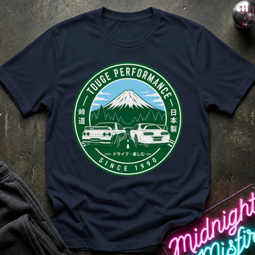 Navy short-sleeve graphic T-shirt laid flat on a dark textured surface. The shirt features a large circular green and white print with a mountain, two cars, and the text 'TOUGE PERFORMANCE' and 'SINCE 1990'.