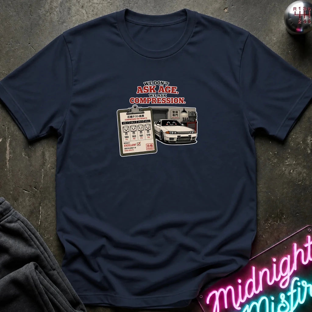 A navy blue short-sleeve graphic t-shirt laid flat on a dark textured surface. The graphic features the text 'WE DON'T ASK AGE, WE ASK COMPRESSION.