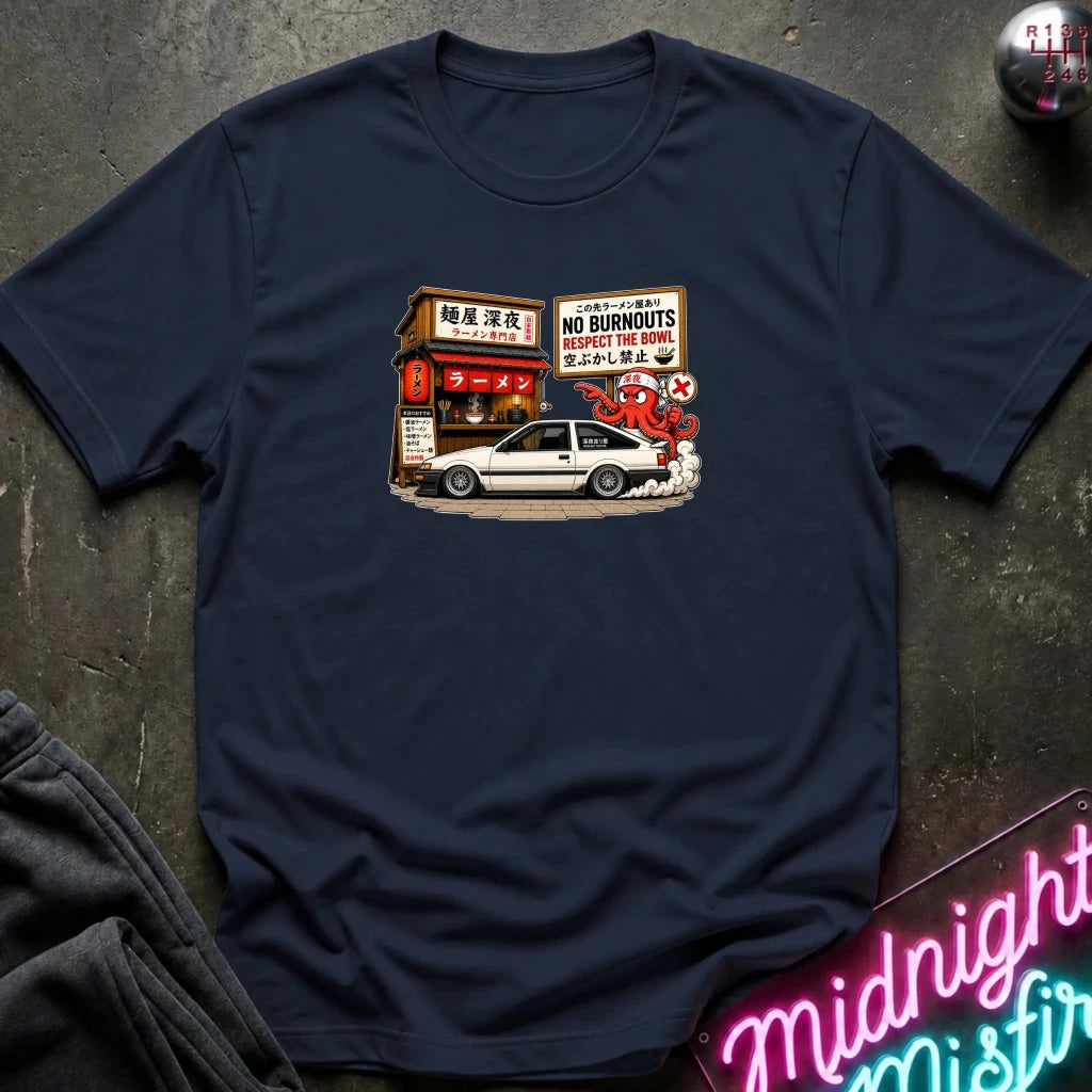 A navy short-sleeve t-shirt laid flat on a dark textured surface, featuring a graphic print of a white sports car parked in front of a Japanese street shop with signs reading 'NO BURNOUTS'.