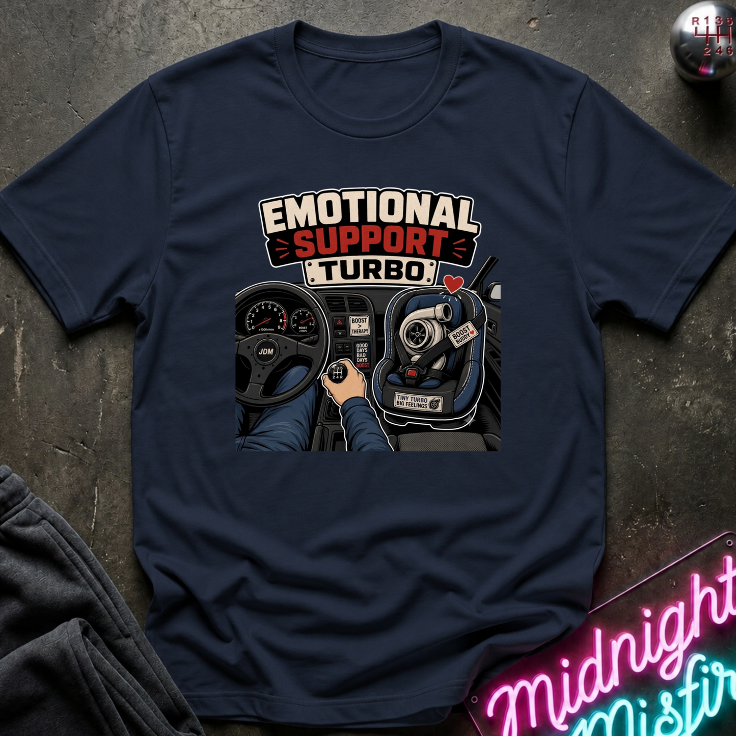 A navy blue graphic t-shirt laid flat on a dark textured surface. The shirt features a graphic print of a car's dashboard and steering wheel with the text 'EMOTIONAL SUPPORT TURBO' in white and red.