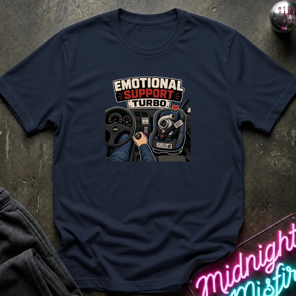 A navy blue graphic t-shirt laid flat on a dark textured surface. The graphic features the words 'EMOTIONAL SUPPORT TURBO' above an illustration of a car dashboard and steering wheel with a hand on the wheel.