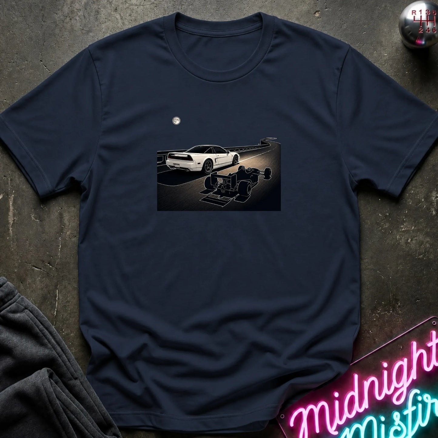 A navy blue short-sleeve crewneck t-shirt laid flat on a dark textured surface, featuring a graphic print of a white sports car and a shadow car on a night road scene. The shirt is styled with dark gray sweatpants, a silver ball, and a neon sign in the corners.