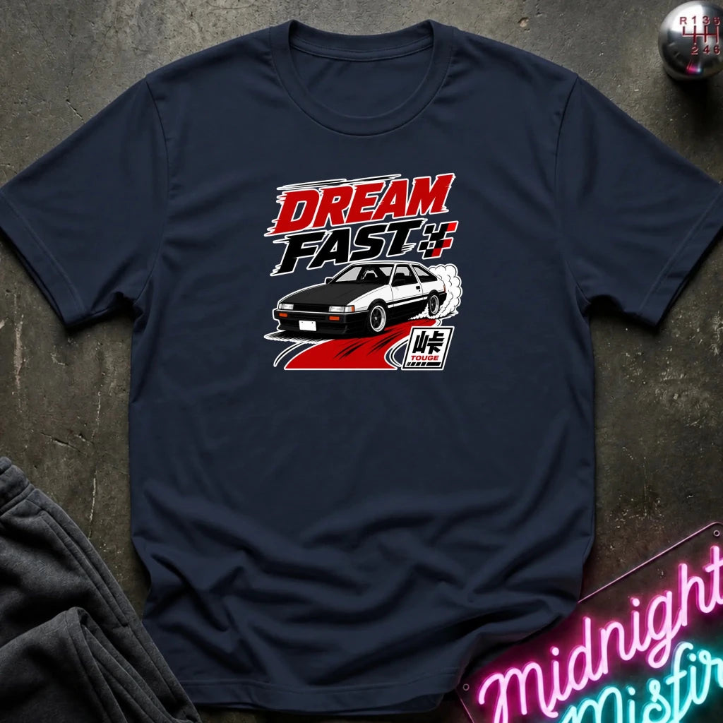 A navy blue short-sleeve graphic T-shirt laid flat on a dark textured surface. The shirt features a red, black, and white 'DREAM FAST' graphic with a car illustration.