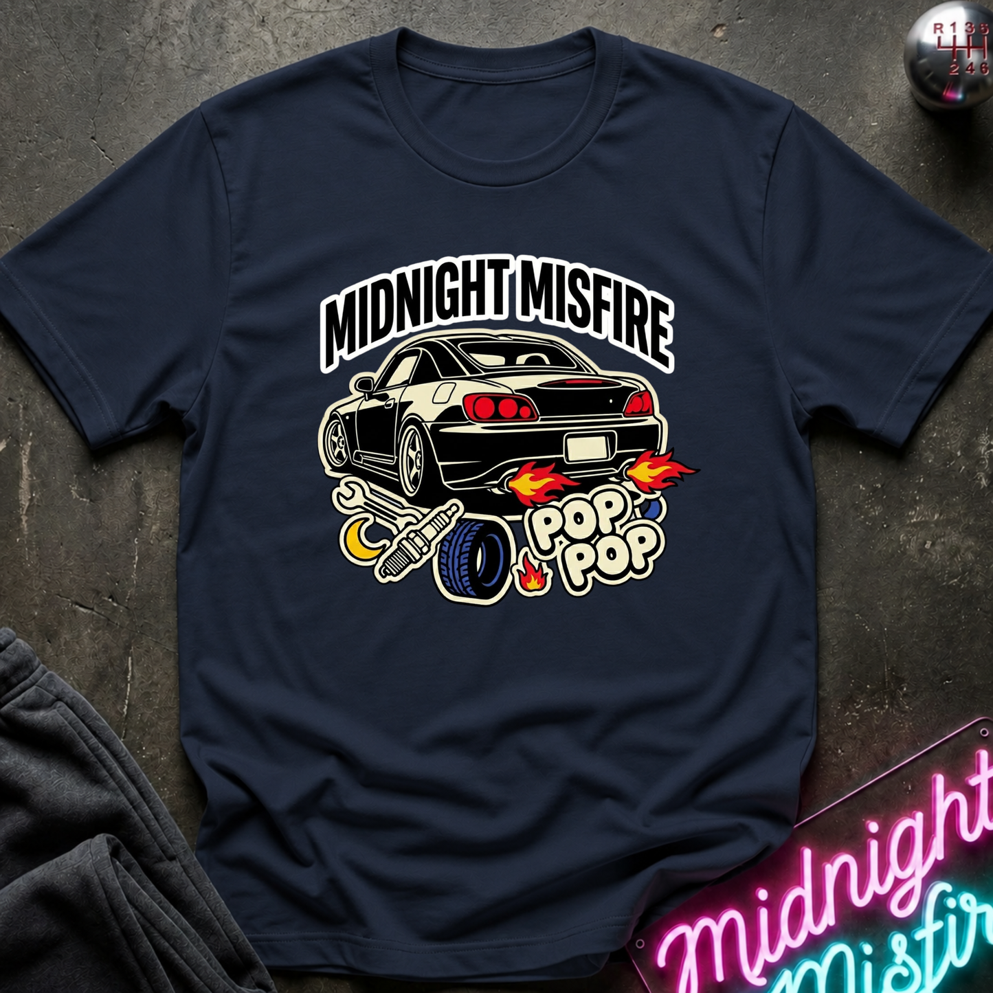 A navy blue crewneck t-shirt laid flat on a dark textured surface, featuring a graphic print of a black sports car with flames and the text 'MIDNIGHT MISFIRE' and 'POP POP'.